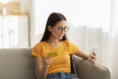 Gözlüklü genç bir kadın evdeki kanepesinde oturuyor, akıllı telefonunu ve kredi kartını online alışveriş için kullanırken gülümsüyor. Doğal ışık odayı dolduruyor, rahat bir atmosfer yaratıyor..