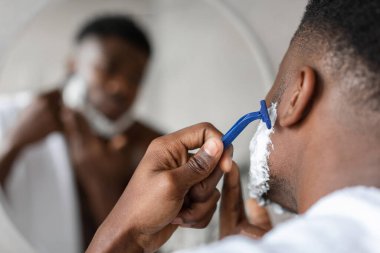 Modern bir banyoda, siyah bir adam aynanın önünde duruyor, yüzünü tıraş etmek için bir tıraş bıçağı kullanıyor. Traş kremi sürerken işine odaklanıyor, dingin bir atmosfer yaratıyor..