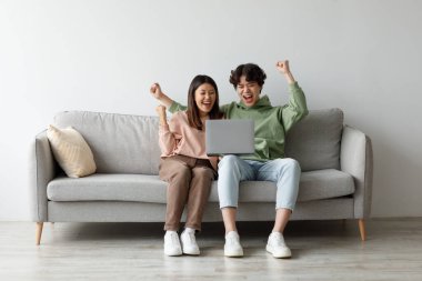 Genç bir Asyalı çift evdeki bir kanepede otururken online iş başarılarını sevinçle kutluyor. Dizüstü bilgisayarlarını kullanırken gülümsüyorlar ve evet diyorlar..
