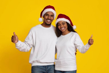 Neşeli bir çift, parlak sarı bir arka plana karşı poz veriyor. İkisi de Noel Baba şapkası ve beyaz kazak giyiyor. Gülümsüyorlar ve başparmaklarını kaldırıyorlar, Noel 'in neşeli ruhunu somutlaştırıyorlar..