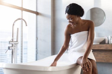 Gülümseyen çekici siyah bayan modern bir banyoda zarif bir küvetin kenarında oturuyor. Havluya sarılıp sabah rutininin tadını çıkarırken su sıcaklığını kontrol ediyor..