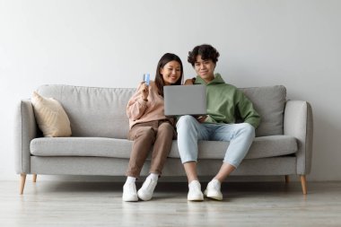 Genç bir Asyalı çift online alışveriş yapmak için dizüstü bilgisayar ve kredi kartı kullanırken koltuklarında dinleniyor. Samimi oturma odalarındaki online alışveriş deneyimleriyle mutlu ve meşgul görünüyorlar..