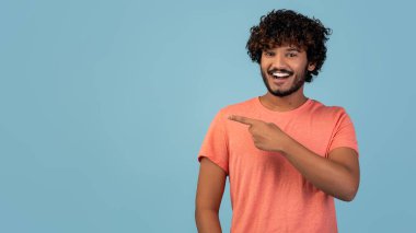 Kıvırcık saçlı ve sakallı genç Hintli bir adam boşluğu işaret ederken gülümsüyor. Pembe bir tişört giyiyor ve mavi bir stüdyo geçmişine karşı poz veriyor. Reklam için mükemmel..