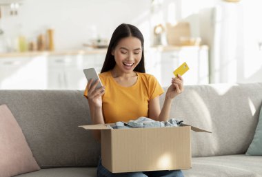 Mutlu bir kadın kanepeye oturur ve hevesle içi kıyafet dolu bir kutuyu açar. Bir elinde cep telefonu, diğer elinde kredi kartı var. Belli ki online alışveriş deneyiminden zevk alıyor..