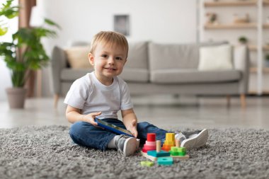 Neşeli genç bir çocuk yumuşak bir halıya oturur, elinde tablet tutarken renkli yapı taşlarıyla meşgul olur. Doğal ışık, modern yaşam alanını yakınlardaki bitkilerle doldurur..