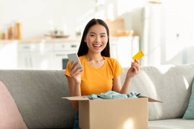 Heyecanlı bir Asyalı kadın oturma odasında bir koltukta oturuyor, bir elinde kredi kartı, diğer elinde akıllı telefon var. Eşyalarla dolu bir karton kutuyu açarken gülümsüyor..