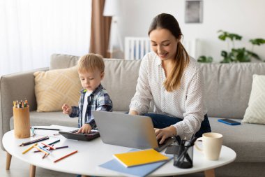 A mother is focused on her laptop while sitting at a coffee table in a bright living room. Her young son plays with a calculator beside her, surrounded by art supplies.