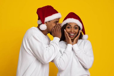 Bir adam ona fısıldarken gülümseyen bir kadın şaşırır. İkisi de Noel Baba şapkası ve beyaz kazak giyiyor. Parlak sarı arka plan, neşeli bir bayram anını yakalayarak şenlik havasına katıyor..