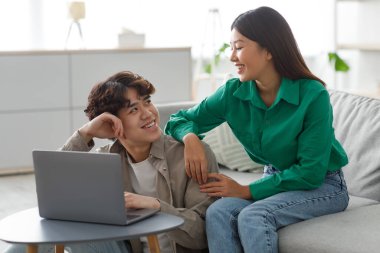 Genç Asyalı çift rahat oturma odalarında dizüstü bilgisayarlarıyla internette gezinmeyi seviyor. En sevdikleri online aktivitelere katılırken gülücüklerini ve kahkahalarını paylaşıyorlar..