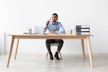 Arap bir adam evdeki bir ofiste masasında oturuyor ve bir dizüstü bilgisayarla çalışıyor. Düşünceli görünüyor, bir kalem tutuyor ve görevlerini düşünürken gözlerini kaçırıyor. Bu onun iş gününde sakin ve ciddi bir an..