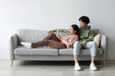 Young Asian couple enjoys quality time on a comfortable couch at home, happily browsing a smartphone and exploring the internet. They share a relaxing moment, engaged with each other.