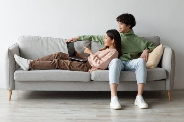 A loving young Asian couple cuddles on a comfortable couch while watching a movie on their laptop. They laugh and engage in an online video chat, creating a cozy atmosphere at home.