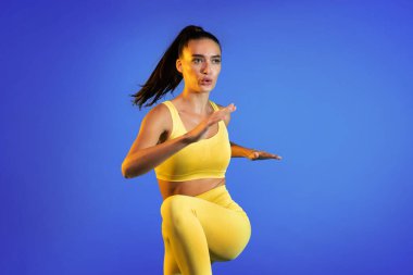 Bir kadın parlak sarı aktif giysi giyerek dinamik bir egzersiz yapıyor. Katı mavi bir zemine karşı hareket halinde, egzersiz yaparken odak ve atletizm gösteriyor..