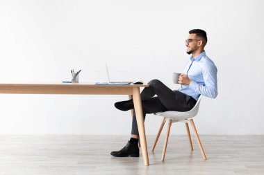 Genç bir Arap, parlak bir odada bir masada oturuyor, bilgisayarını kullanarak gülümsüyor. Elinde bir fincan sıcak kahve tutuyor. Keyifli bir an yaşıyor ve rahatlatıcı bir atmosfer yaratıyor..