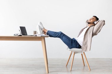 Huzurlu bir ortamda masasında oturan, kablosuz kulaklık takan ve dinlendirici müzik dinleyen sakin bir adam. Sırtını masaya yaslar ve rahatlarken gülümser..