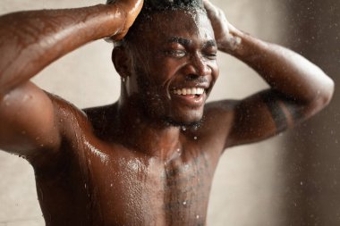 Siyah bir erkek banyo banyosunda duruyor, gözleri kapalı ve saçlarını sıcak suyun altında yıkarken gülümsüyor. Sahne, kapalı bir ortamda rahatlama ve kişisel bakıma vurgu yapıyor..