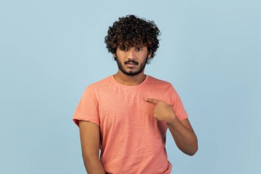 Kıvırcık sakallı Hintli bir adam kendini işaret ederken şaşırmış görünüyor. İfadesi, kendisine gönderme yapılıp yapılmadığını kontrol ederken merak ve kafa karışıklığını gösteriyor. Arka plan pürüzsüz bir mavi..