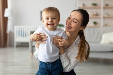 Bir anne, küçük oğlunu sıcak bir oturma odasında tutarken gülümser. Güneş ışığı boşluğu doldurur, oyun zamanı için sıcak bir atmosfer yaratır ve bağ kurar..