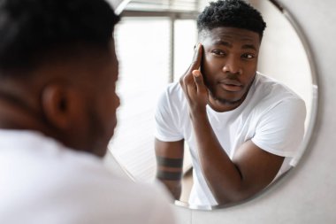 Tıraşsız bir siyah adam banyo aynasında yansımasına bakarken yüzüne dokunuyor. Tıraş rutinine hazırlanıyor. Bu an kendine bakmanın ve tımar etmenin görüntüsü..