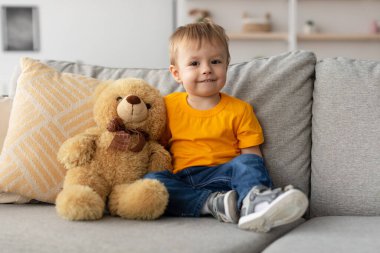 Sarı gömlek giyen neşeli genç bir çocuk, yumuşak bir oyuncak ayının yanında gri bir kanepede otururken gülümsüyor. Rahat atmosferde ışık dekoru var..