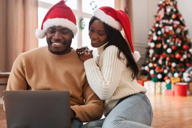 Neşeli siyah bir çift modern bir dairede yerde oturuyor, kırmızı Noel Baba şapkaları takıyorlar. İnternetten alışveriş yapmak ya da Noel arifesinde birlikte Noel filmleri izlemek için dizüstü bilgisayar kullanıyorlar..