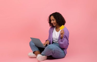 Genç siyahi bir kadın dizüstü bilgisayarıyla yerde oturuyor ve kredi kartını online alışveriş için kullanıyor. Parlak pembe bir stüdyoda, eğlenceli ve modern bir hava sergiliyor..