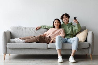 A young Asian couple relaxes on their comfortable sofa, enjoying a movie night at home. They switch channels with a remote control, sharing laughter and moments together.