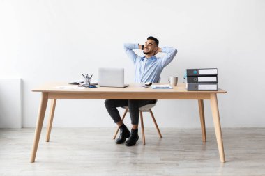 Neşeli bir Arap, modern bir ofis masasında elleri başının arkasında oturur ve rahat bir sandalyeye yaslanır..