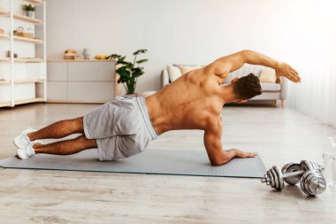 Bir adam, parlak, ferah bir kapalı alanda yan tahta egzersiziyle meşgul olur. Yoga paspası kullanıyor. Yanında da halterler var..