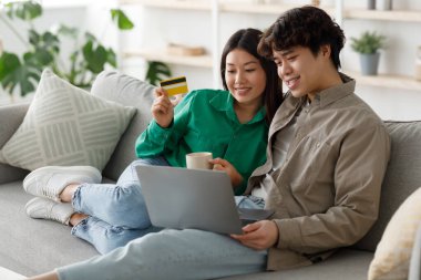 Neşeli bir Asyalı çift kanepelerinde oturup bilgisayarla internetten alışveriş yapıyor. Ortaklardan biri kredi kartını tutarken diğeri de evde alışveriş yaparken kahvesini yudumluyor..