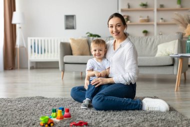 Neşeli bir anne, her ikisi de gülümseyen küçük çocuğuyla birlikte halıya oturur. Renkli oyuncaklar yakındaki sıcak, davetkar bir oturma odasına saçıldı..