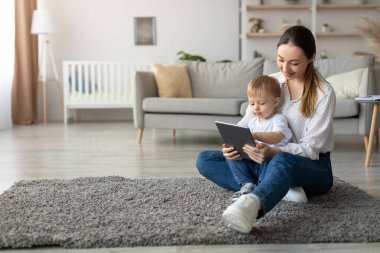 Bir anne, her ikisi de tablet ekranına odaklanmış küçük çocuğuyla yerde oturuyor. Konforlu oturma odasında yumuşak bir halı, yeşil bir kanepe ve pencerelerden gelen doğal bir ışık var..