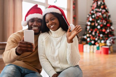 Her ikisi de kırmızı Noel Baba şapkası takan neşeli siyah bir çift yerde oturmuş sevdiklerine video araması yapıyorlar. Oturma odası Noel için güzel süslenmiş bir ağaç ve hediyelerle..