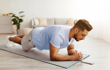 Bir adam parlak bir odada egzersiz paspası üzerinde tahta egzersizi yapıyor. Odaklanmış görünüyor ve telefonunu egzersiz için kontrol ediyor, formunu koruyor..