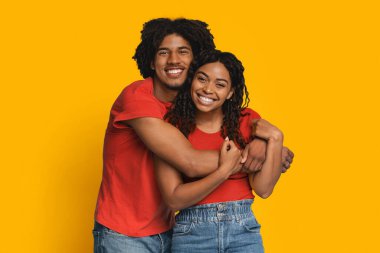 Mutlu bir çift birbirine sarılıyor, ikisi de kırmızı bluz ve kot pantolon giyiyor. Parlak sarı bir arkaplan önünde büyük bir gülümseme sergiliyorlar..