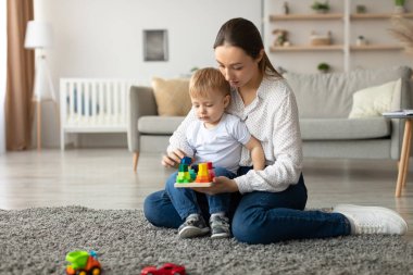 Bir anne ve küçük çocuğu, rahat oturma odalarında yumuşak bir halının üzerinde renkli yapı taşlarıyla eğlenceli bir aktiviteye girişiyor. Hava sıcak ve davetkar..