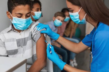 Modern bir klinikte, bir sağlık çalışanı Afrikalı Amerikalı bir çocuğa Covid-19 aşısı yapıyor. Arka planda birkaç çocuk daha bekliyor. Güvenlik için maske takıyorlar..
