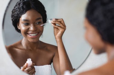 Güzel, Afro-Amerikalı bir bayan gözlerinin altına yüz serumu sürerken kendi yansımasına gülümsüyor. Günlük güzellik rutininin bir parçası olarak cildinin pürüzsüzlüğünün tadını çıkarıyor..