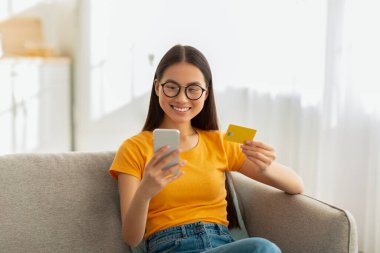 Genç Asyalı bayan evdeki bir koltukta rahatça oturuyor, akıllı telefonunu ve kredi kartını kullanarak internetten alışveriş yaparken gülümsüyor, uzaktan kumandalı bankacılığın rahatlığının keyfini çıkarıyor..