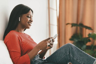 Bir kadın akıllı telefonunda mesaj atarken gülümseyerek kanepede dinlenir. Kulaklık takıyor ve doğal ışığı olan rahat ve sıcak bir yaşam alanıyla çevrili..