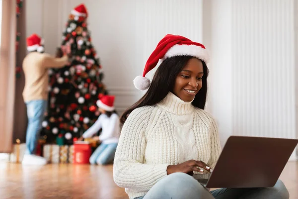 Kırmızı Noel Baba şapkalı neşeli Afro-Amerikalı kadın hediye almak için dizüstü bilgisayar kullanarak yerde oturuyor. Arka planda, aile üyeleri bir Noel ağacı süsleyerek şenlikli bir ortam yaratıyorlar..