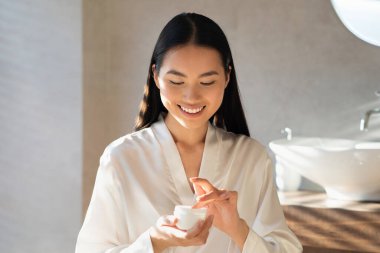 Beyaz bornozlu gülümseyen Asyalı genç bayan parlak banyosunda bir kavanoz güzellik ürünü tutuyor. Sabah rutininden zevk alır. Güneş ışığı alan güzel cildini aydınlatır..