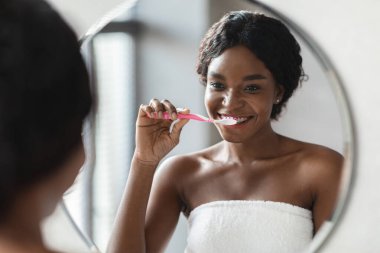 Diş telleri olan mutlu siyah kadın banyo aynasının önünde dikilirken dişlerini fırçalar. Üstü başı havluyla kaplı ve sabah diş bakımı rutininin tadını çıkarıyor..