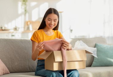 Genç bir kadın kanepesinde oturuyor, neşeyle yeni kıyafetlerle dolu bir kutu açıyor. Teslimatta gülümsüyor, sıcak oturma odasında alışveriş deneyiminin tadını çıkarıyor..