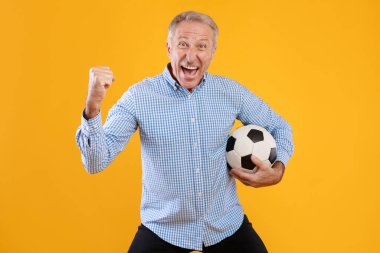 Olgun bir adam bir futbol topuyla sevinçle poz verir, kazanan bir jest yapar. Canlı sarı bir arkaplanda duruyor, bir golü heyecanla kutluyor. Spor hevesini yakalamak için mükemmel..