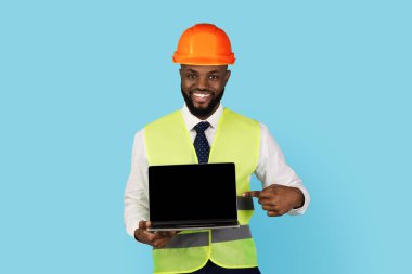 Gülen Siyah İnşaat İşçisi elinde boş ekranla dizüstü bilgisayarı işaret ediyor. Sert şapka ve güvenlik yeleği giyen yakışıklı Afro-Amerikan yapıcı web sitesi için fotokopi alanı gösteriyor.