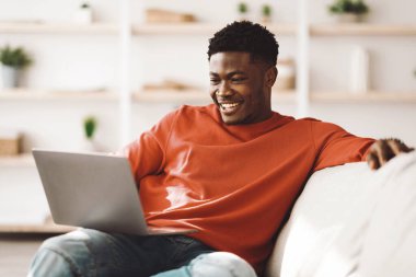 Genç bir adam kanepeye rahatça oturur, dizüstü bilgisayarıyla uğraşırken gülümser. Konforlu yaşam alanı hafif dekor ve bitkiler içeriyor, rahatlamak için rahatlatıcı bir atmosfer yaratıyor..