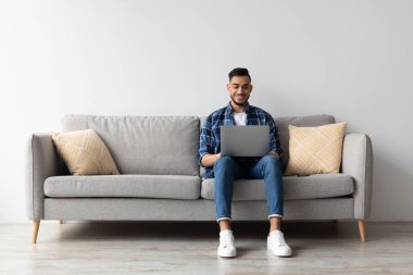 Neşeli bir Arap evdeki kanepede oturur ve uzak işi için dizüstü bilgisayar kullanır. Rahatlamış görünüyor ve uzaktan çalışmanın esnekliğinden zevk alıyor. Etrafı rahat bir oturma odasıyla çevrili..