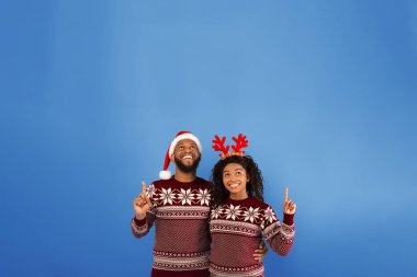 Tatil teklifi. Birebir aynı Noel süveterleri içinde mutlu siyah eşler fotokopi alanına bakıyor geyik boynuzu takan kadın ve mavi arka planda duran Noel Baba şapkalı adam.
