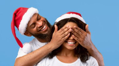 Heyecanlı afro-amerikan adam gözlerini kapatıyor, şaşırmış genç kadın kollarını yana açıyor ve gülümsüyor, Sants şapkası takan çift, sarı stüdyo arka planında birlikte duruyorlar.